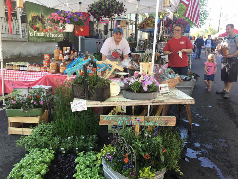 Two people smiling and standing behind a Red Top Farm Market booth stocked with local favorites, herbs, and flowers.