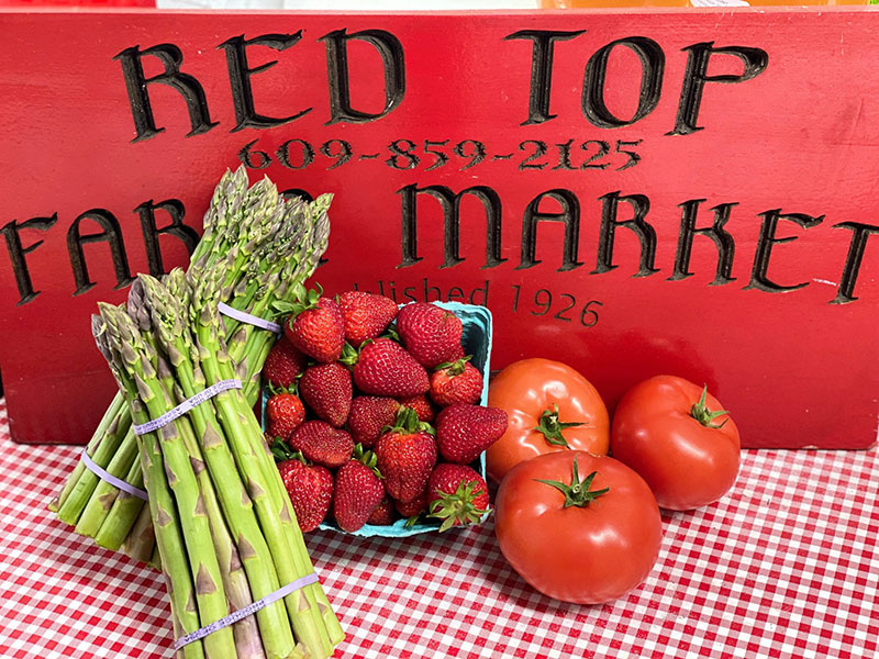 A red sign saying "Red Top Farm Market" sitting on a red gingham table cloth next to asparagus, strawberries, and tomatoes.