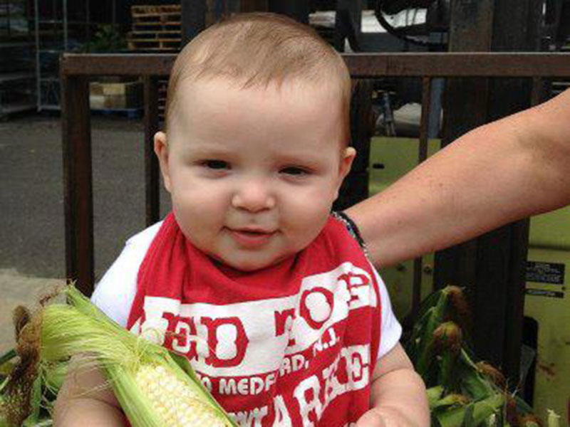 A baby wearing a Red Top Farm Market bib smiling at the camera while holding an ear of corn.